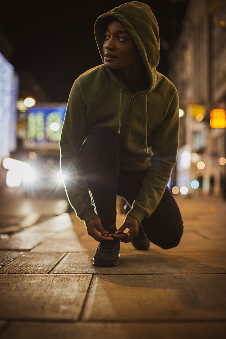 Young female runner tying shoelace on city sidewalk at night
