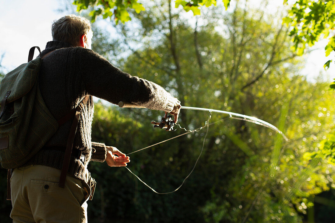 Man with backpack casting fly fishing pole below sunny trees