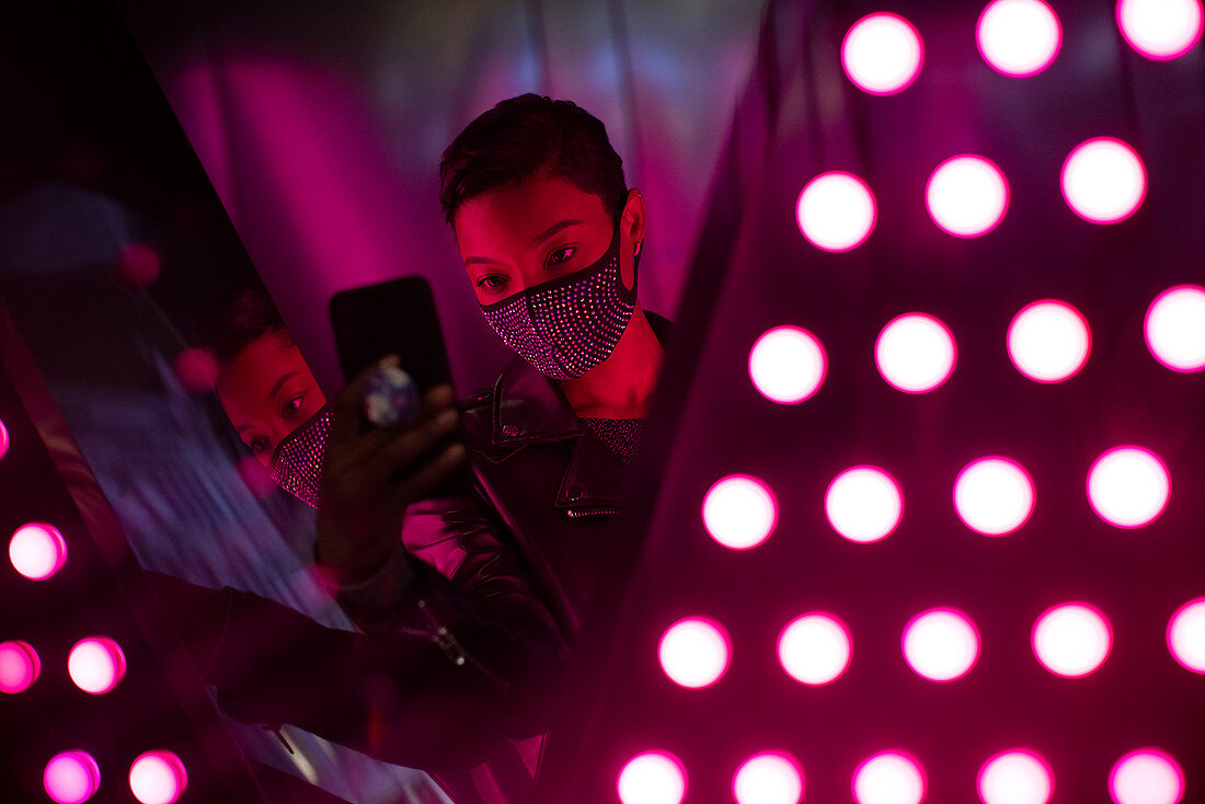 Young woman in face mask taking selfie at mirrored light