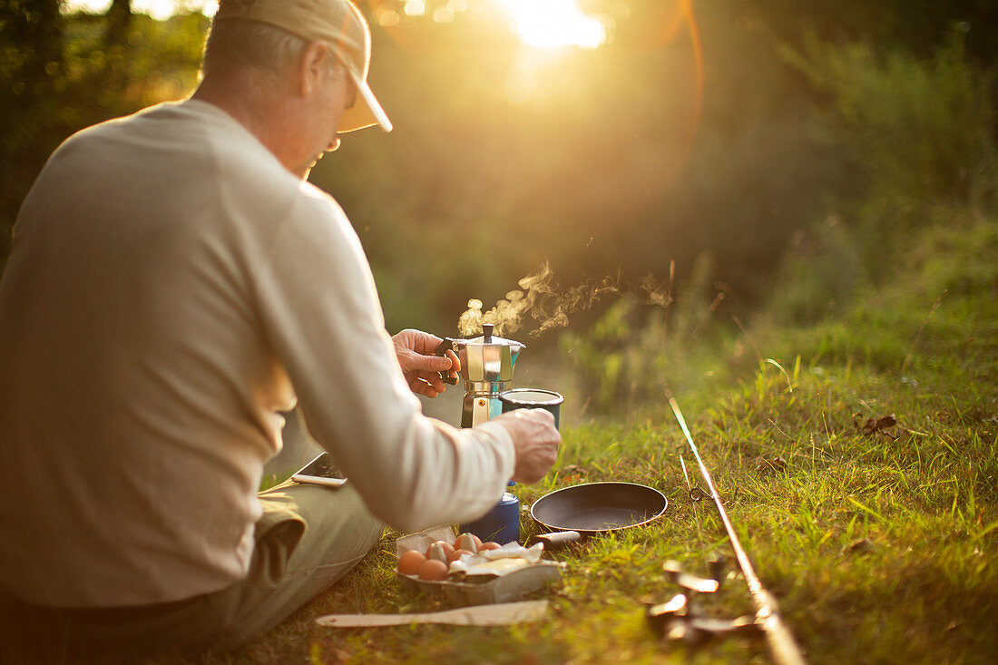 Man making coffee on riverbank during break from fly fishing