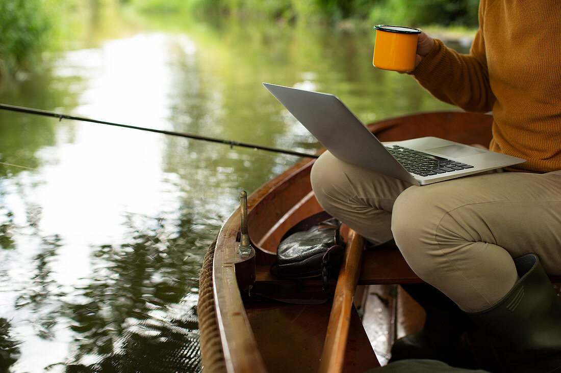 Man fly fishing and using laptop with coffee in a boat