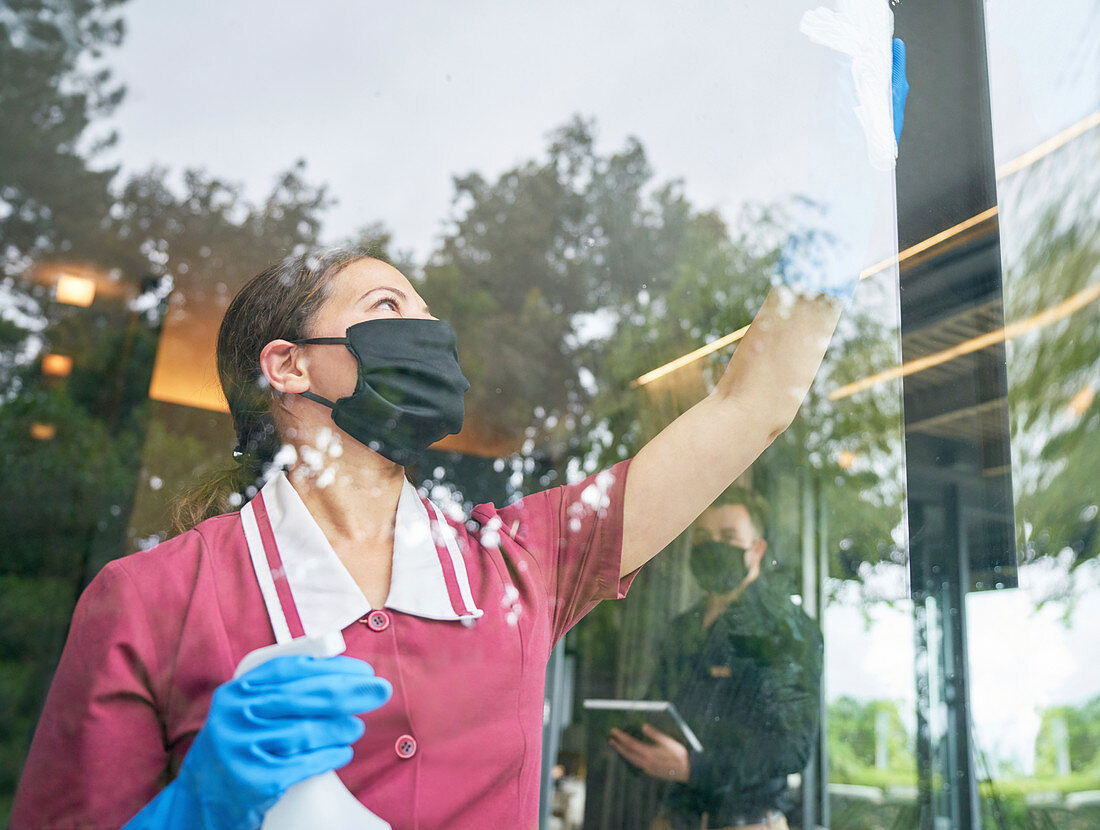 Female hotel maid in face mask cleaning hotel window