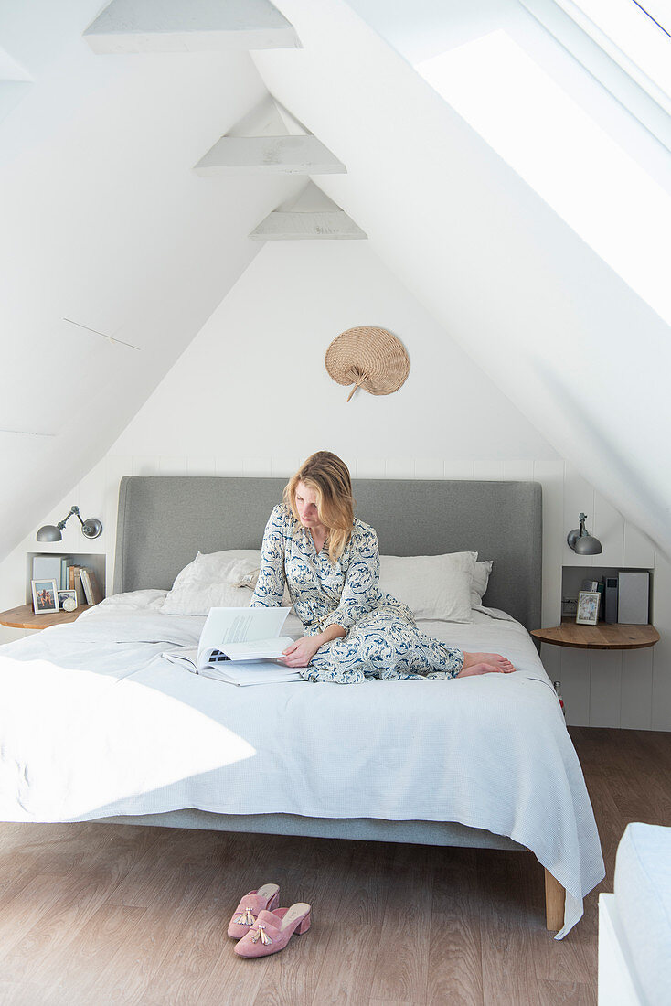 Blonde Frau sitzt mit Buch auf Bett im Dachgeschoß