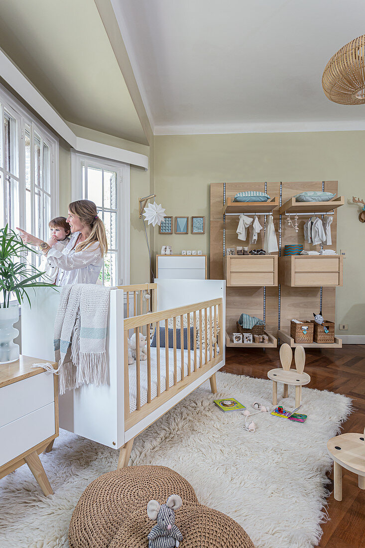 Mutter mit Kleinkind am Fenster im Kinderzimmer mit Gitterbett auf Hochflorteppich