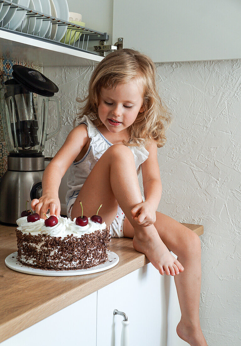 Little girl sitting on the kitchen counter and tasting cherry cake with her finger