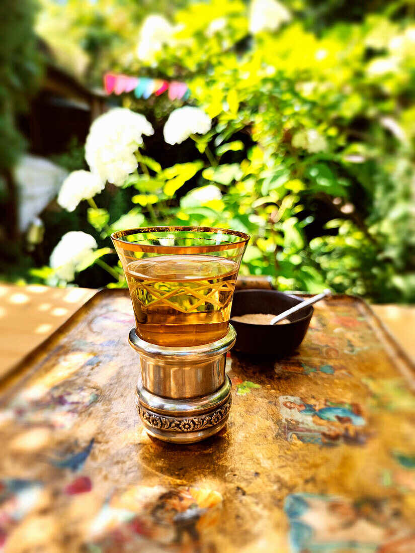 Ein Glas Minztee und ein Schälchen brauner Zucker auf Tablett im Garten