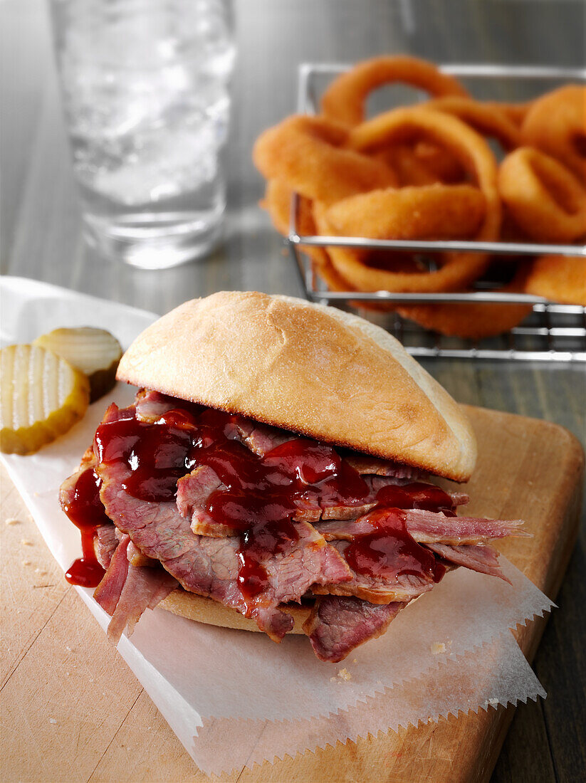 A beef brisket sandwich with BBQ sauce on a bun with pickles and fried onion rings