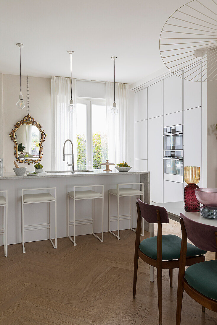 White kitchen counter with bar stools and gold-framed mirror, dining table with chairs in the foreground