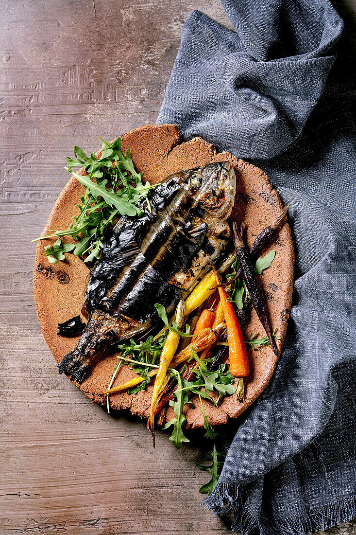 Grilled cooked fresh gutted sea bream on ceramic plate wrapped in bamboo leaves served with herbs, colorful carrots
