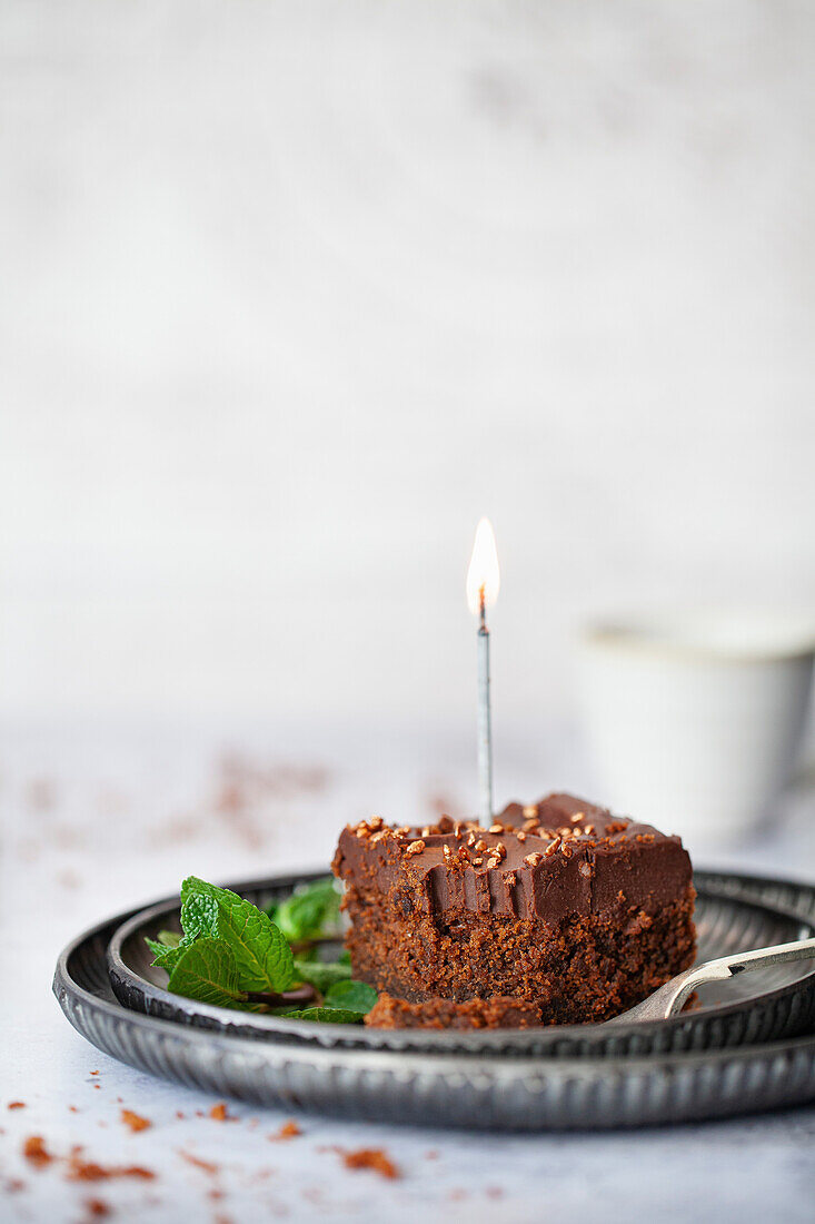A Square of Chocolate cake topped with ganache, bronze sprinkles and a lit candle