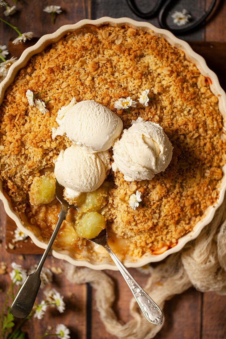 A part eaten gooseberry crumble with ice cream on top and spoons in the baking dish