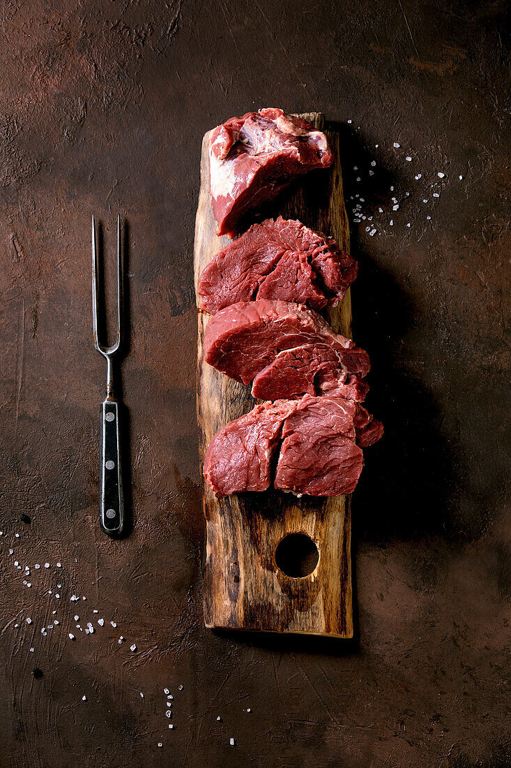 Sliced raw beef tenderloin meat for steaks on wooden board with metal meat fork