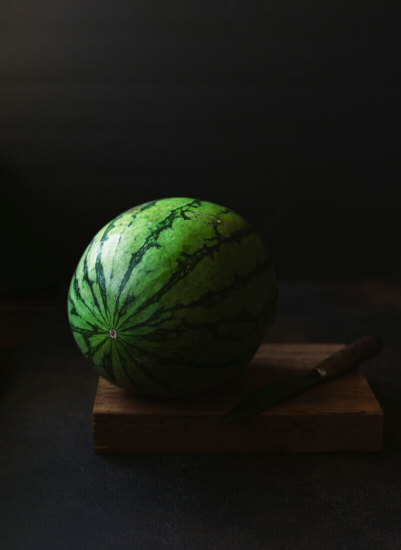 Ganze Wassermelone mit Messer auf Holzbrett vor schwarzem Hintergrund