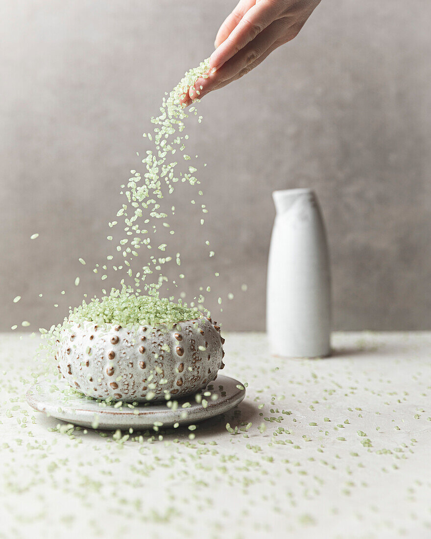 A hand pours green bamboo-infused rice into a decorative ceramic bowl