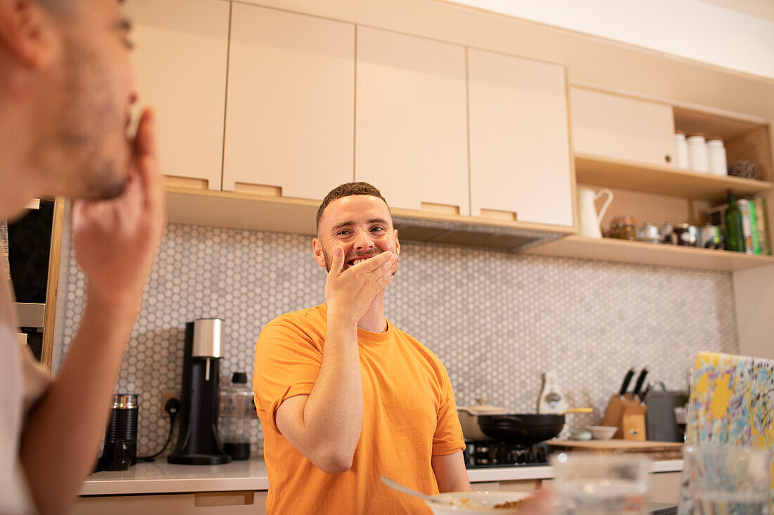 Happy gay male couple laughing and eating in kitchen
