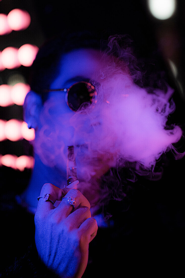 Young man with vape pen blowing smoke in neon light