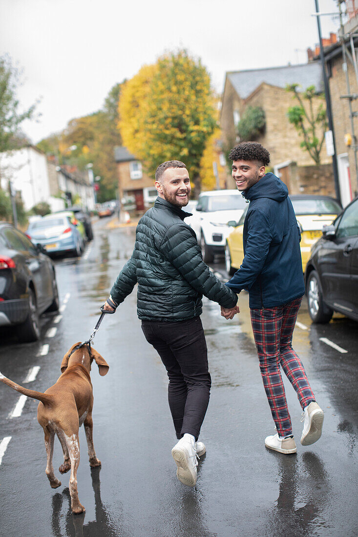 Happy gay male couple walking dog on wet autumn road