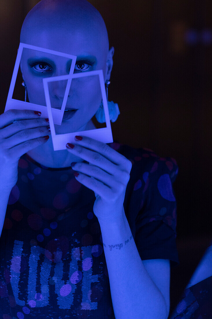 Woman with shaved head holding polaroids in blue light