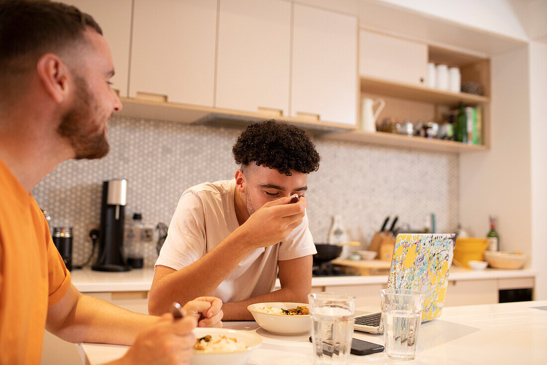 Gay male couple eating and using laptop at kitchen counter