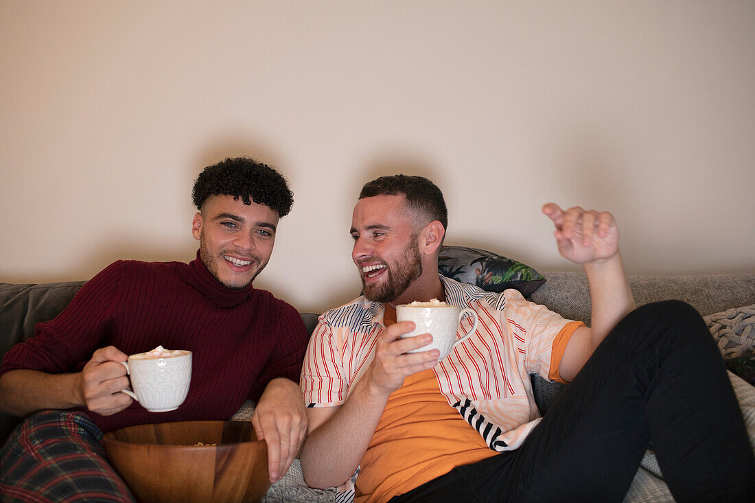 Happy gay male couple drinking hot cocoa watching TV on sofa