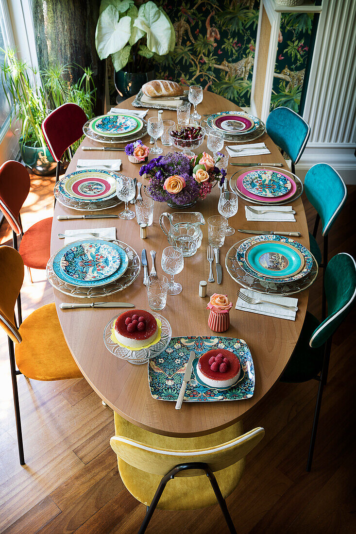 Ovaler, gedeckter Esstisch mit bunten Tellern, Kristallgläsern und Blumenstrauß