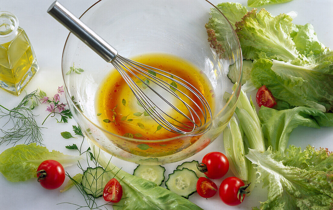 Glass bowl with red wine vinegar-olive oil vinaigrette and a whisk, with salad ingredients next to it