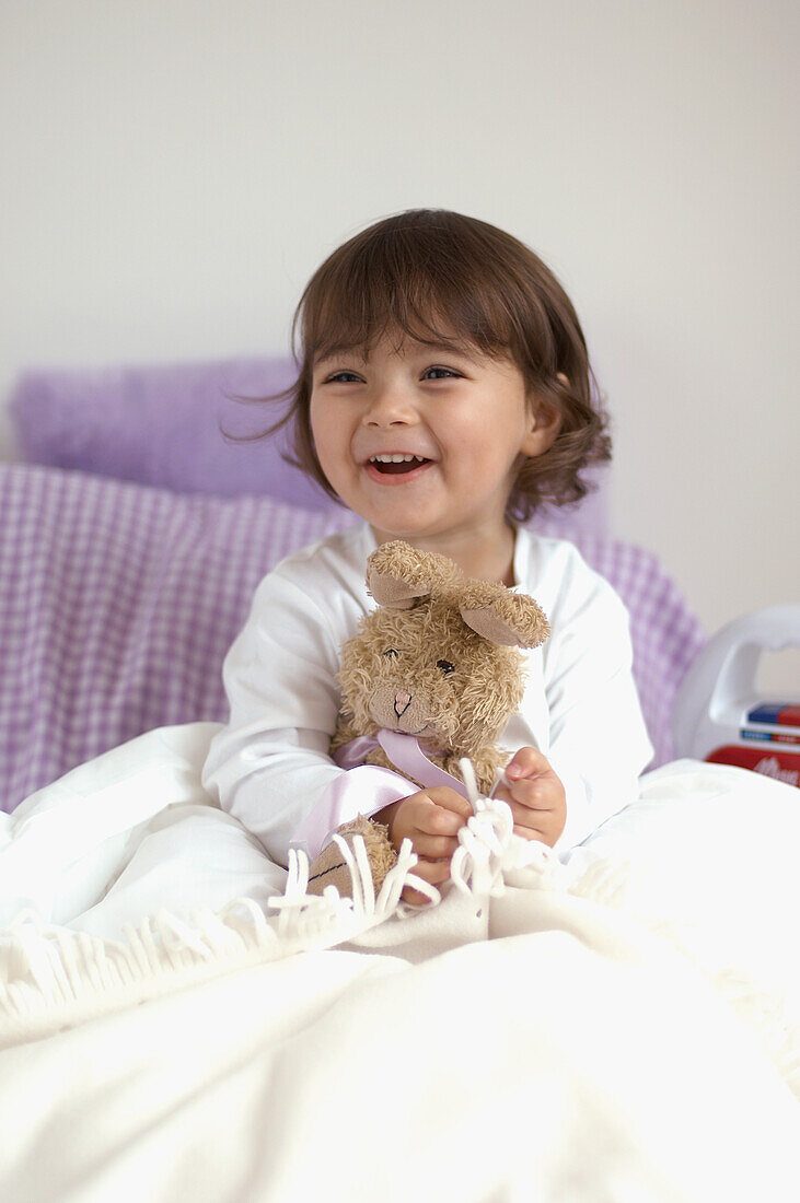 Young girl in pyjamas sitting up in bed, holding toy rabbit