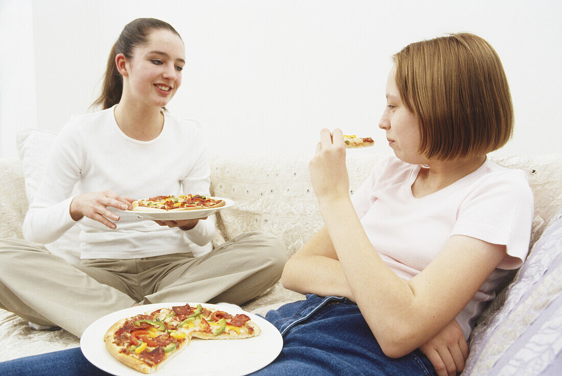Two teenage girls sitting on a sofa eating pizza