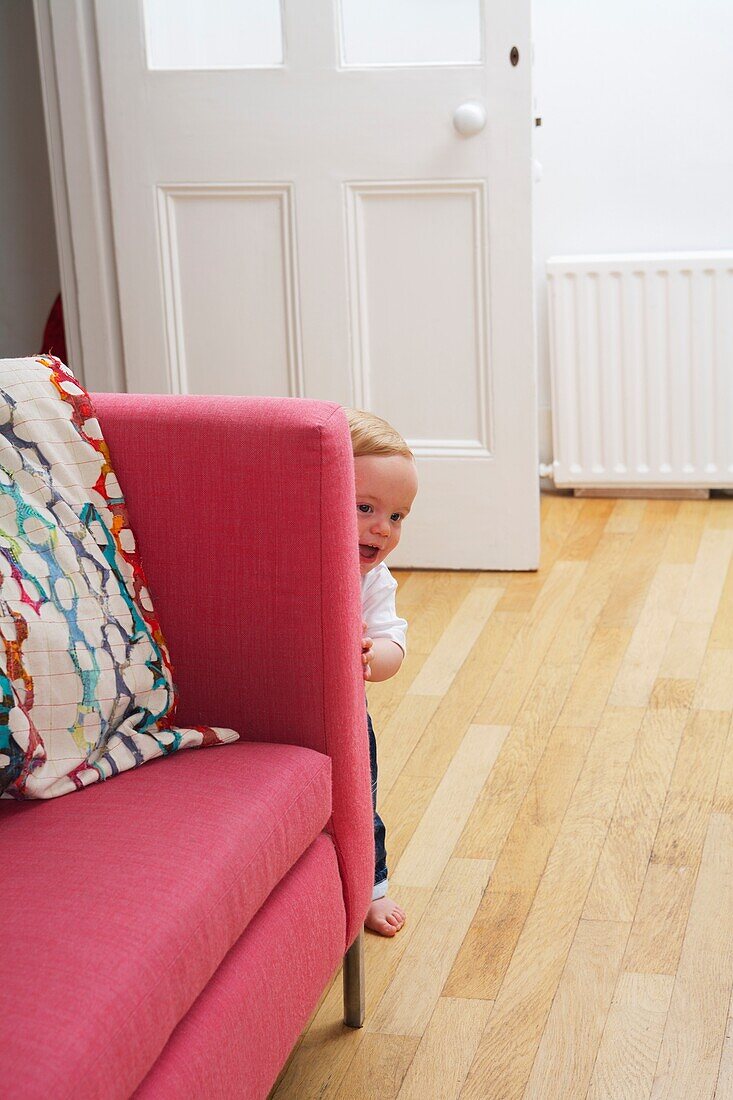 1 year old baby boy peeking around side of sofa