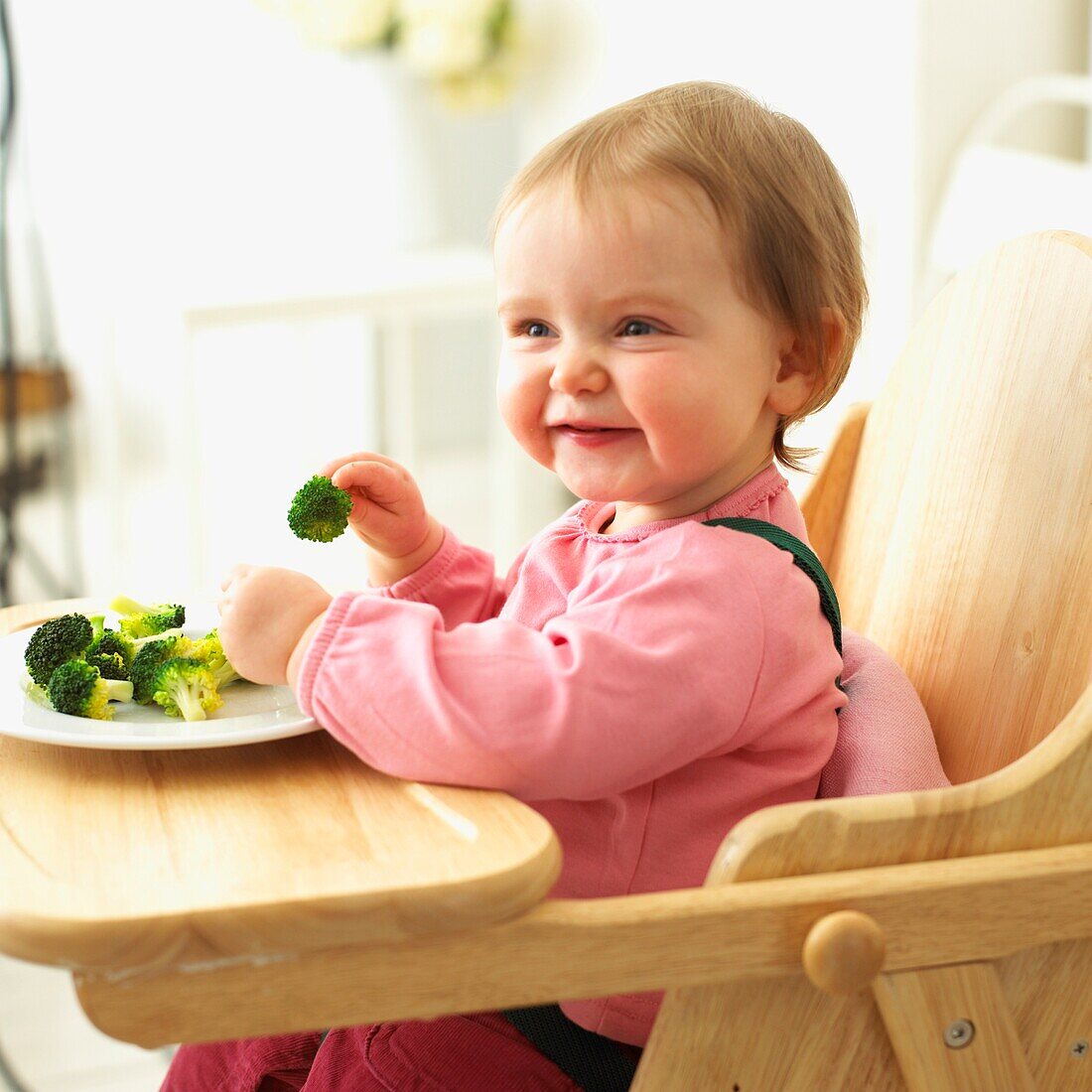 Baby girl sitting in wooden high chair eating broccoli