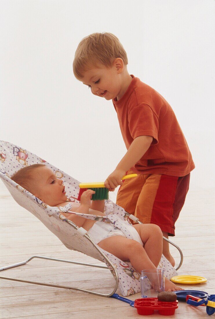 Young boy brushing baby who is sitting in bouncing cradle