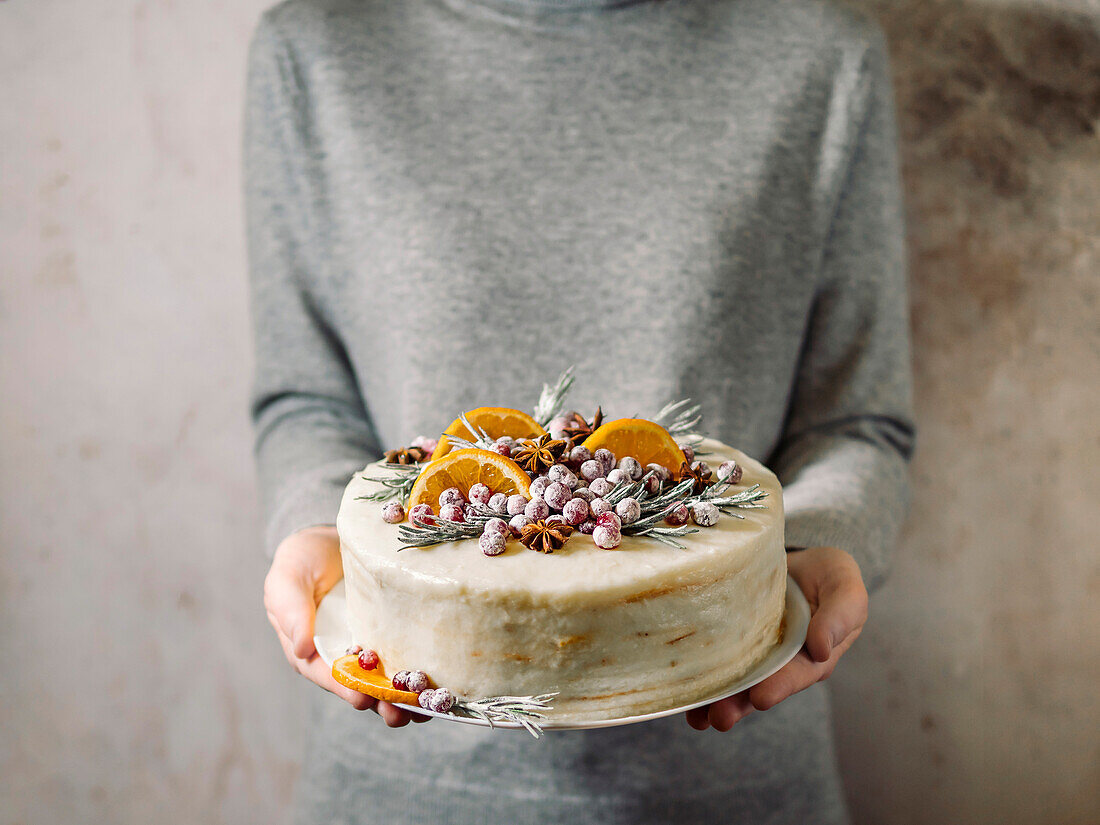 Hands hold white cream christmas cake decored rosemary, cranberries and oranges slices