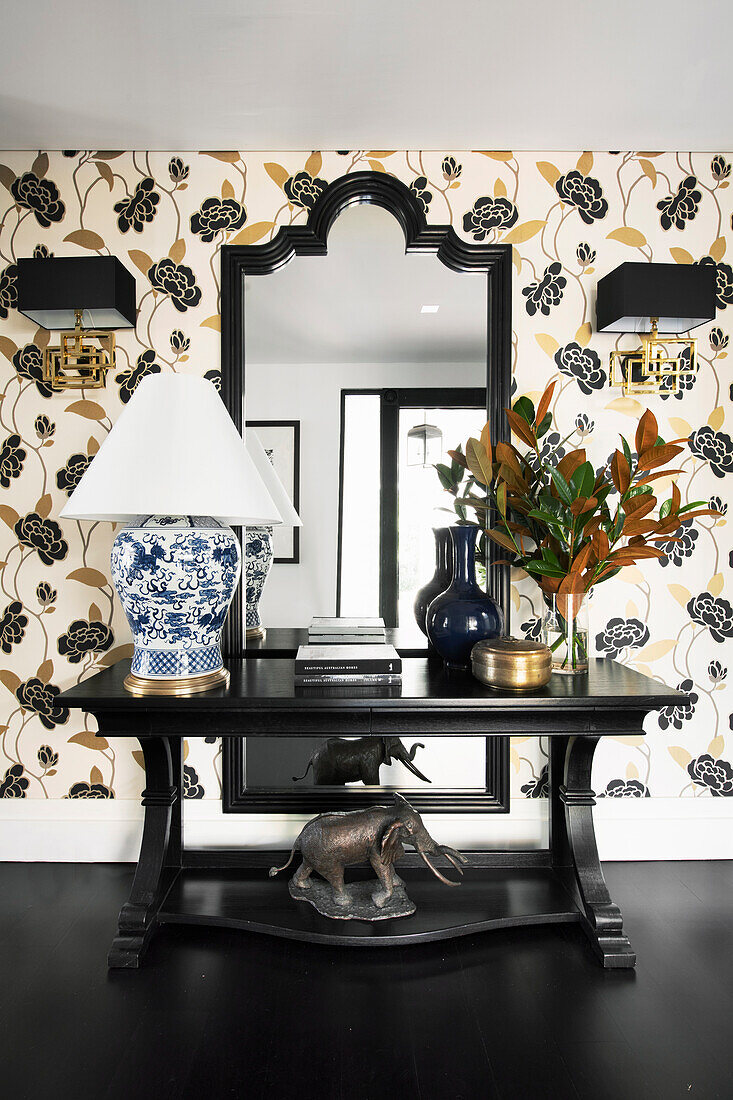 Black console table with table lamp and bouquet of flowers in front of wallpaper with floral design and wall mirror