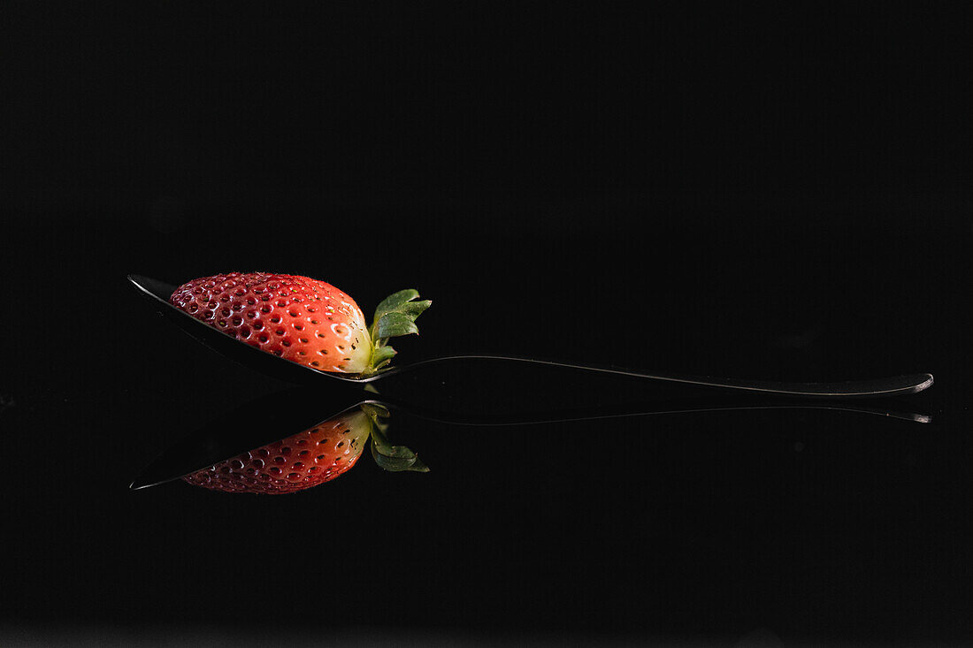 Half a strawberry cut and on a spoon, black reflective background