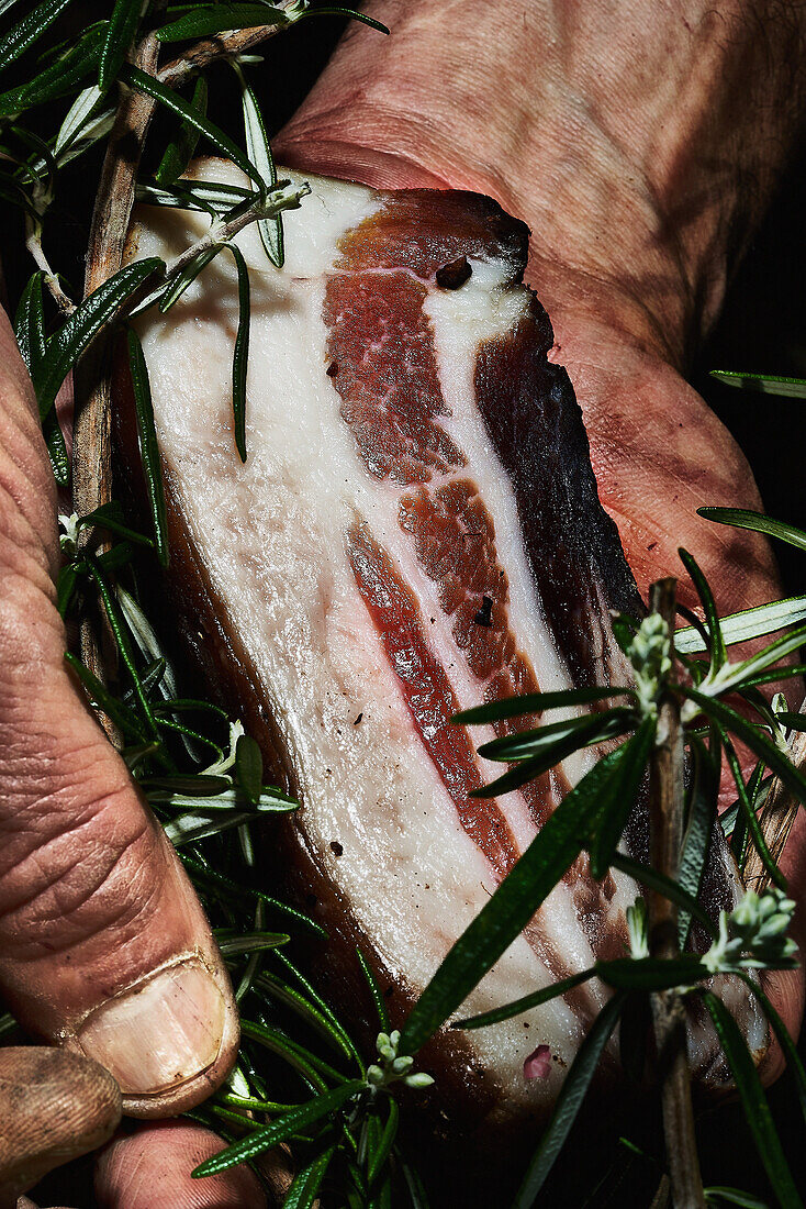 Hände halten ein Stück geräucherten Speck mit Rosmarin