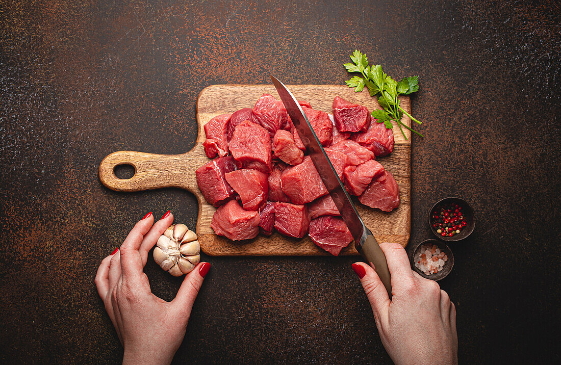 Female hands chopping raw beef meat in cubes on wooden cutting board for cooking stew