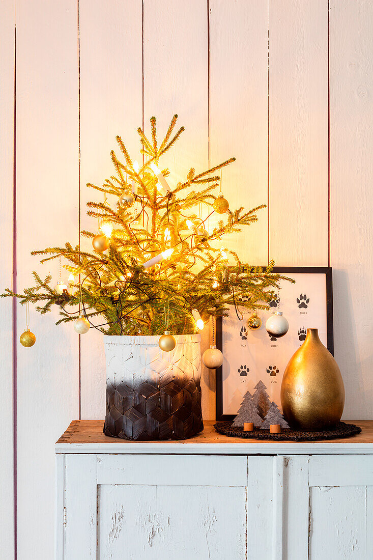 Weihnachtliche Dekoration mit beleuchteter Mini-Tanne im Topf, goldener Vase und Bild auf Kommode