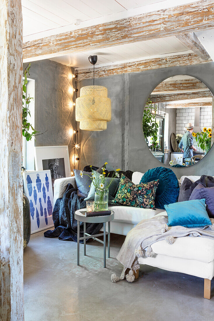 Old sofa with decorative cushions under round mirror in open living room with concrete floor and wooden beams