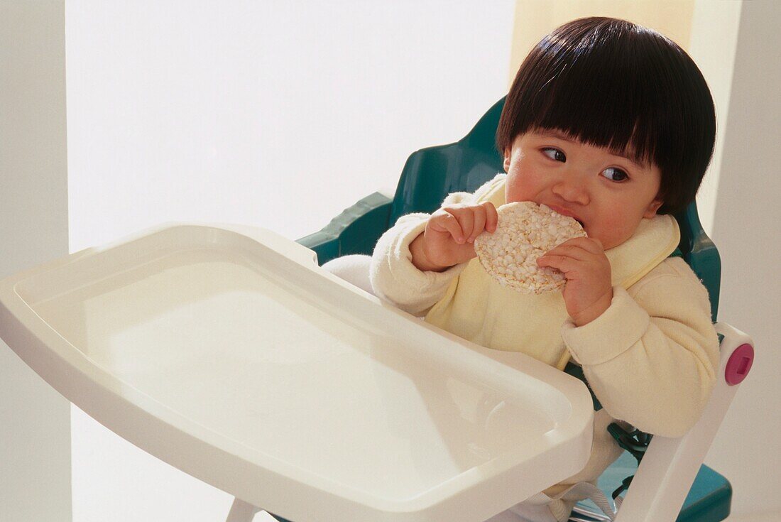 Toddler sitting in high chair feeding herself a rice cake