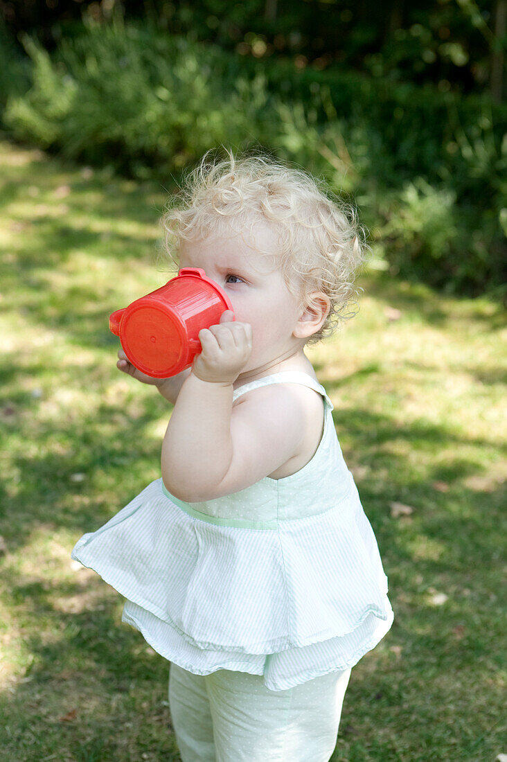 Baby girl in a garden drinking from red plastic beaker
