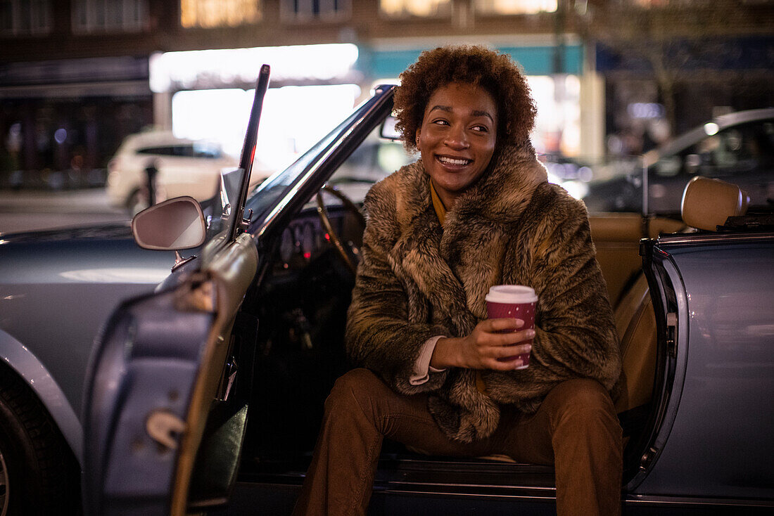 Happy young woman with coffee at convertible at night