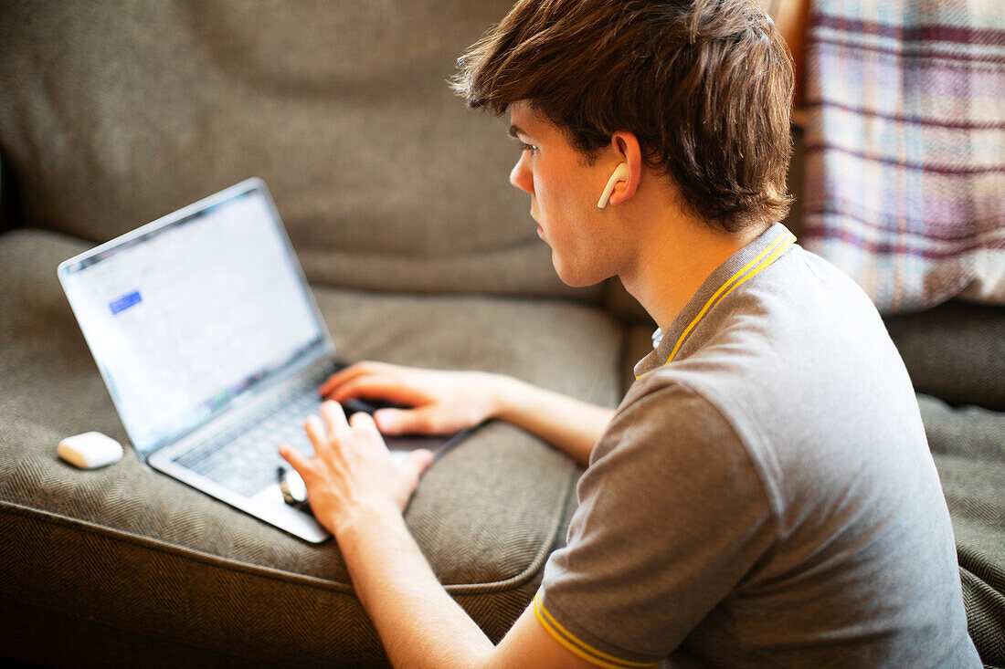Teenage boy with ear buds using laptop on sofa at home