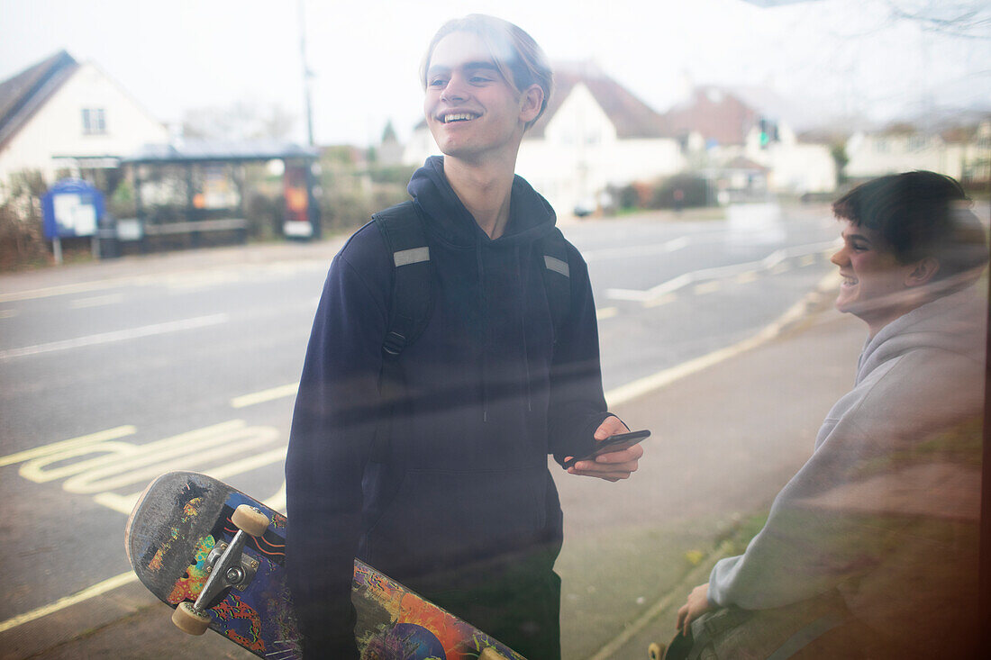 Happy teenage boy with smartphone and skateboard at bus stop
