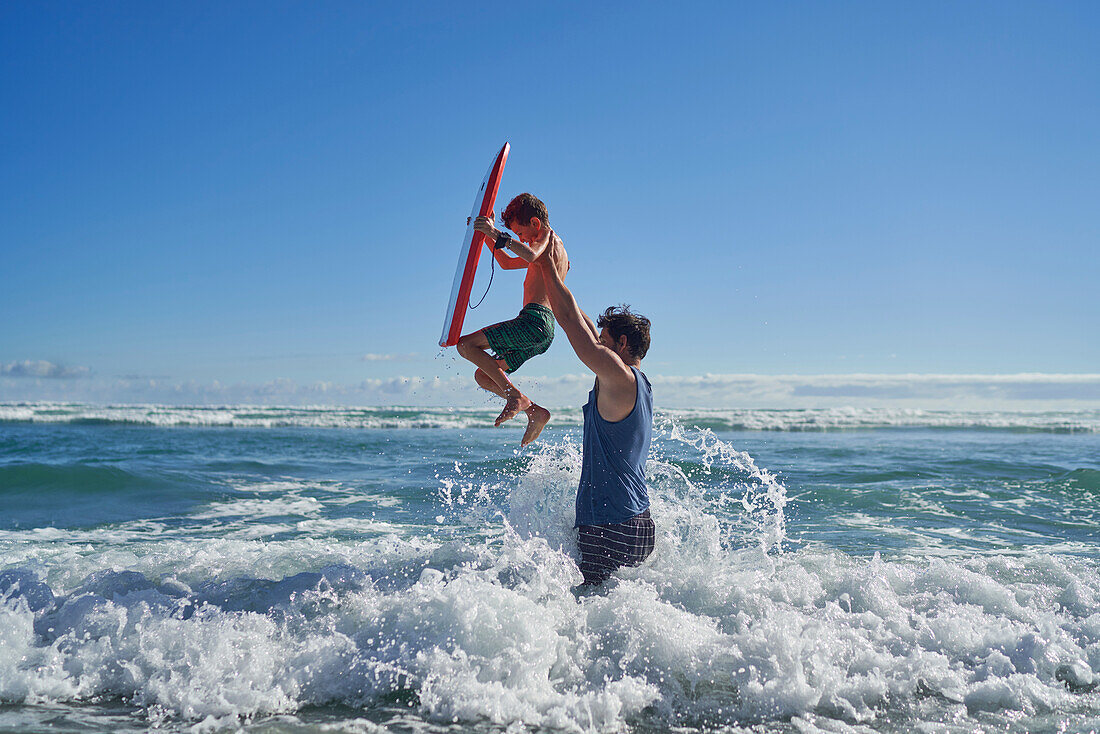 Father lifting son with body board above sunny ocean wave