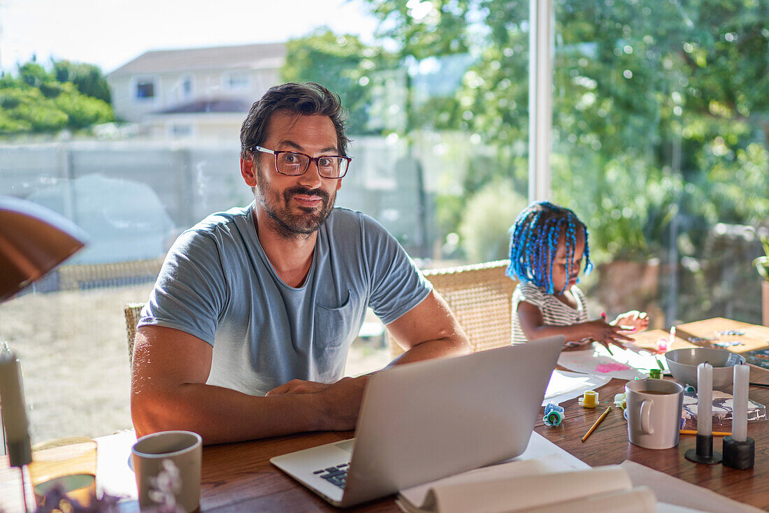 Father working at laptop next to daughter finger painting