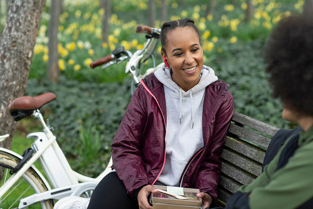 Happy young woman eating lunch with friend on park bench