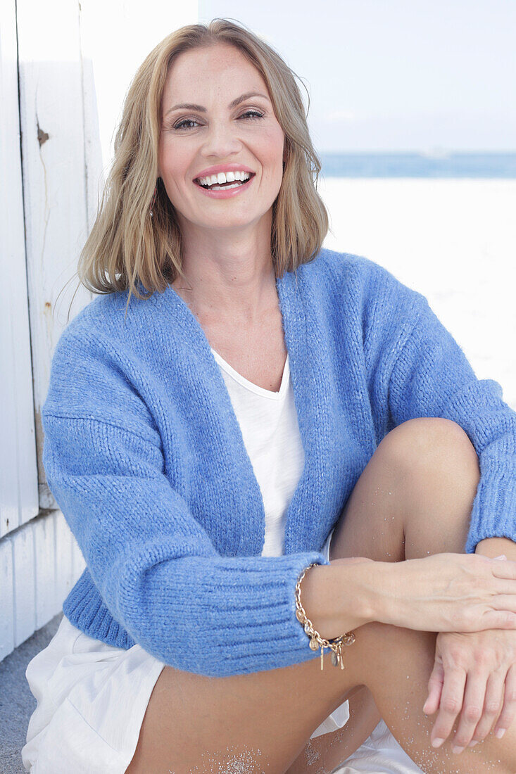 Long haired woman in white summer dress and blue cardigan sitting on the beach