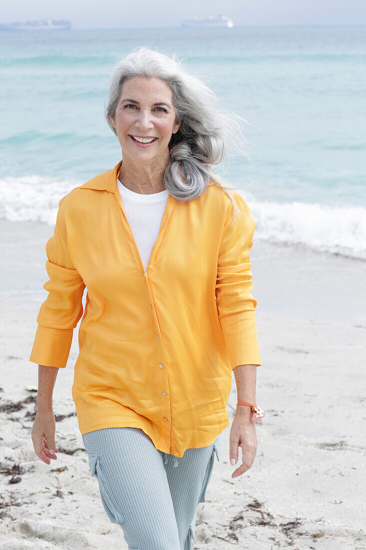 Reife Frau mit grauen Haaren in orangefarbener Bluse und Hose am Strand