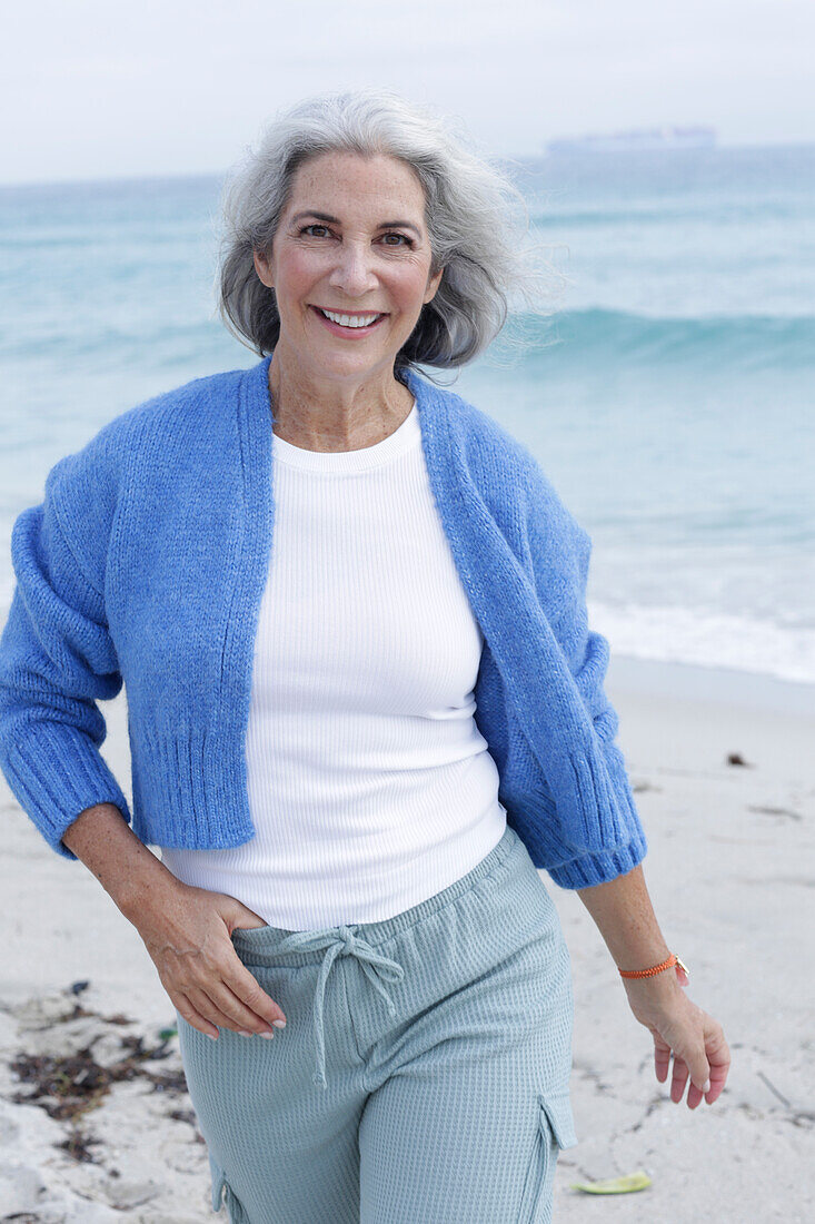 Reife Frau mit grauen Haaren in weißem T-Shirt, blauer Strickjacke und Hose am Strand