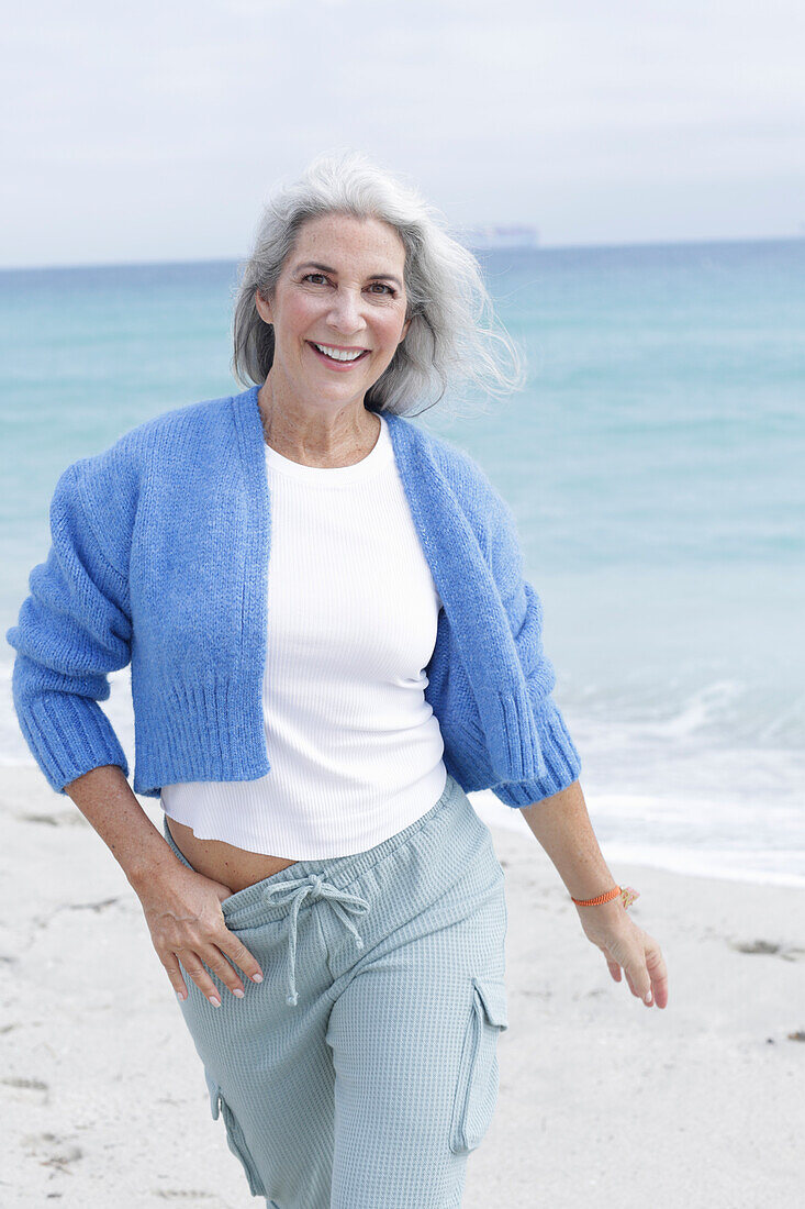 Mature woman with grey hair in white t-shirt, blue cardigan and trousers on the beach